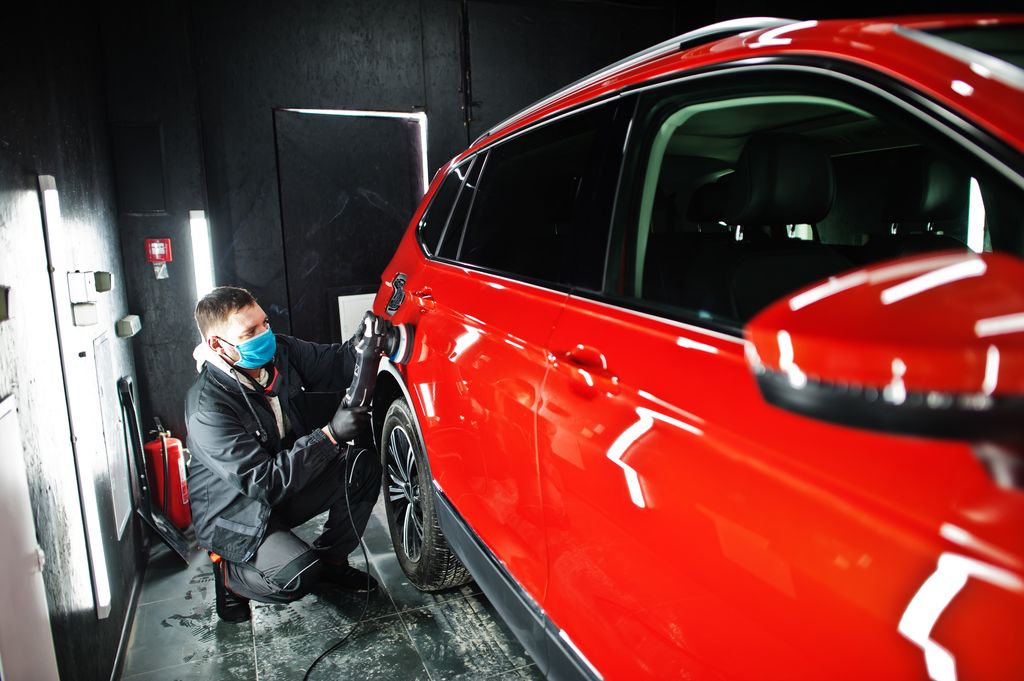 A technician wearing a protective face mask uses an orbital polisher to refine the paint on a red car during a professional ceramic coating preparation.