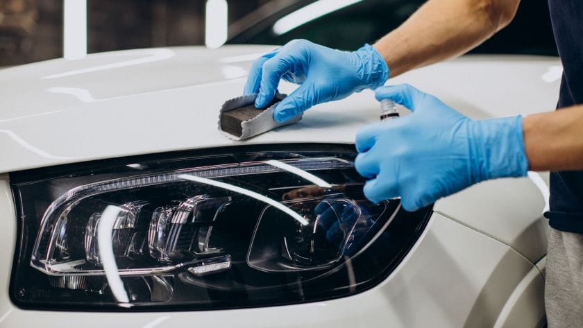 A technician wearing blue protective gloves applying ceramic coating to a white car's front panel and headlight.