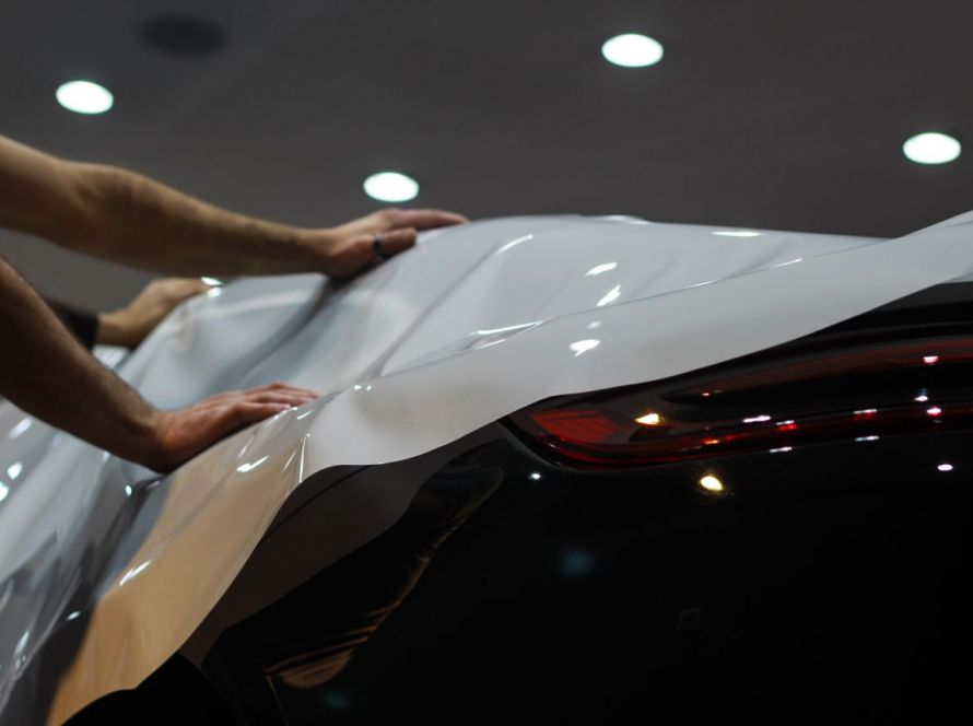 Two technicians carefully applying a white vinyl car wrap onto the hood and side of a modern vehicle in a workshop.